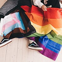 Zu sehen ist eine Regenbogenflagge die über den Beinen von zwei Menschen ausgebreitet liegt.