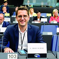 Christian Doleschal sitzt im EU-Parlament an seinem Platz, vor ihm ein Mikrofon und sein Namensschild. Er lächelt in die Kamera.