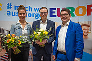 Ilona Schuhmacher, Vizepräsidentin des BJR, Philipp Seitz und Matthias Fack, BJR-Präsident, stehen nebeneinander und lachen in die Kamera. Schuhmacher und Seitz halten Blumensträuße in den Händen.
