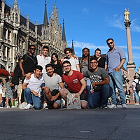 11 internationale Jugendliche stehen zum Gruppenfoto zusammen auf dem Münchner Marienplatz. Sie haben sich gegenseitig die Arme über die Schultern gelegt und lächeln in die Kamera.
