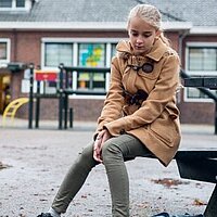 Ein Mädchen sitzt allein auf einer Bank am Spielplatz und schaut traurig.