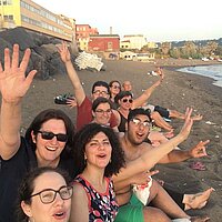 Eine Gruppe sitzt am Strand und hebt die Hände in die Höhe.