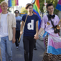 Eine Gruppe junger Menschen läuft Hand in Hand über eine Bücke. Sie tragen Regenbogenfahnen.