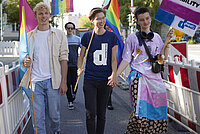 Eine Gruppe junger Menschen läuft Hand in Hand über eine Bücke. Sie tragen Regenbogenfahnen.