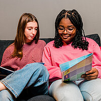 Zwei Mädchen sitzen auf der Couch und schauen sich U18-Infomaterial an. 
