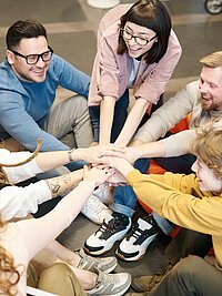 Eine Gruppe von Mitarbeitenden sitzt auf dem Boden im Kreis. Alle halten ihre Hände zu einer motivierenden Geste in die Mitte.
