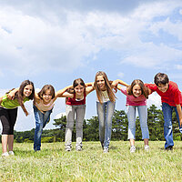 Eine Kindergruppe steht in einer Reihe lachend auf einer Wiede und beugt sich Richtung Kamera