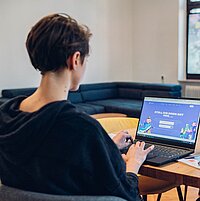 Eine junge Person sitzt an einem Schreibtisch mit dem Rücken zum Betrachter, vor sich einen Laptop und im Hintergrund einen großen Bildschirm