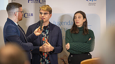 Eine Person in der Mitte spricht in ein Mikrofon, der Moderator und eine weitere Personen stehen links und rechts davon