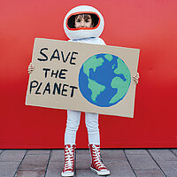 Ein Mädchen in einem Astronautenkostüm mit Helm steht vor rotem Hintergrund und hält ein selbsgebasteltes Schild in den Händen mit einer Weltkugel und dem Text "Save the Planet"