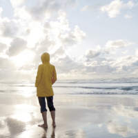 Eine Jugendliche von hinten in gelber Regenjacke mit hochgekrempelten Hosen schaut auf Meer und  Wolken.