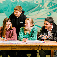fünf Jugendliche stehen um einen Tisch und diskutieren miteinander