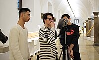 Drei junge Männer machen eine Aufnahme mit IPad in einem Museum und schauen grübelnd auf den Monitor