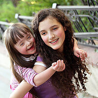 Zwei lachende Mädchen. Ein Mädchen mit Trisomie 21 wird Huckepack getragen.