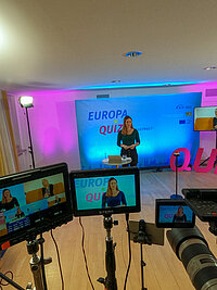 Im Studio steht die Moderatorin. Rechts auf der Leinwand hält die ehemalige bayerische Jugendministerin Trautner eine Videobotschaft. 