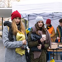Zwei junge warm angezogene Frauen haben Wolldecken in der Hand, dahinter der Ausgabe-Tisch für die Wolldecken.