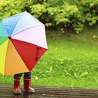 Ein bunt angezogenes Kind in Regenstiefeln und Regenkleidung, Gesicht und Oberkörper sind durch einen bunten Regenschirm verdeckt, der es vor dem Regenbschützt. 