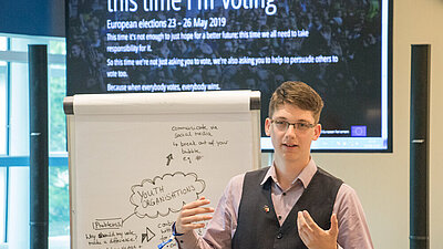 Ein junger Mann steht neben einer Flipchart und präsentiert die Ergebnisse. 