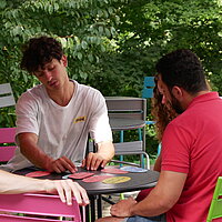 Drei junge Männer und eine junge Frau sitzen an einem Tisch im Freien und diskutieren über bunte Moderationskarten, die vor ihnen auf dem Tisch liegen. 