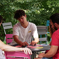 Drei junge Männer und eine junge Frau sitzen an einem Tisch im Freien und diskutieren über bunte Moderationskarten, die vor ihnen auf dem Tisch liegen. 